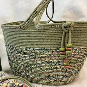 Puede incluir: Un bolso tote de cuerda verde oliva hecho a mano con una sección inferior estampada floral. El bolso presenta un detalle de cuerda anudada con cuentas decorativas y un asa a juego. Se ve un carrete de hilo verde y una tela estampada.