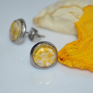 Puede incluir: Un par de pendientes de bot&oacute;n de plata con incrustaciones de resina amarilla y blanca. Los pendientes est&aacute;n sobre una superficie blanca con un p&eacute;talo de flor amarillo.