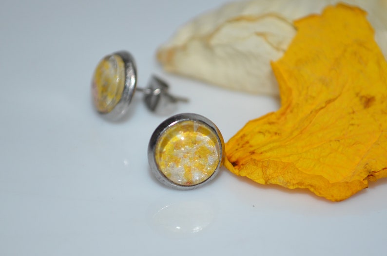 Puede incluir: Un par de pendientes de bot&oacute;n de plata con incrustaciones de resina amarilla y blanca. Los pendientes est&aacute;n sobre una superficie blanca con un p&eacute;talo de flor amarillo.