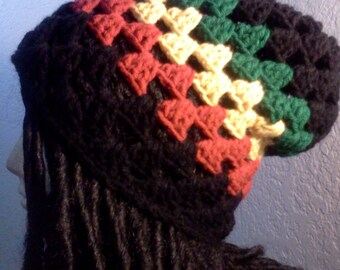 Op de afbeelding: Een zwarte gehaakte beanie met rode, gele en groene strepen. De beanie wordt gedragen door een persoon met lange zwarte dreadlocks.