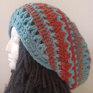 May include: A handmade crochet hat in shades of teal, rust, and brown. The hat features a textured stitch pattern and is designed in a slouchy style. The hat is shown on a mannequin head with dark dreadlocks.