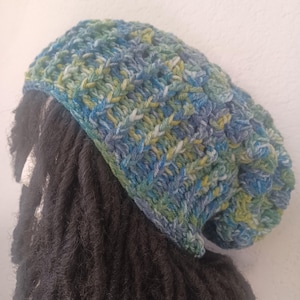 May include: A handmade crochet beanie in shades of blue, green, and yellow. The hat has a textured, woven appearance and a relaxed fit. The beanie is a stylish accessory for cold weather.