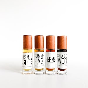 May include: Four small brown glass bottles with labels and black text. The bottles are arranged in a row from left to right. The labels read "Cosmic Lights", "Summer Haze", "Hermetic", and "Shadow Work".