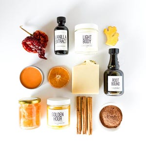 May include: A collection of wellness products including a dried chilli, a bottle of vanilla extract, a jar labelled Light Body, a piece of ginger, an orange slice, a bar of soap, a Root Bound bottle, a jar of honey, a Golden Hour jar, cinnamon sticks, and cocoa.