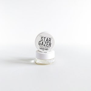 May include: A small, clear glass jar with a white lid, containing a cream product. The lid is labelled "STAR GAZER" in black text. The product is identified as a herbal eye cream. The jar is set against a white background.