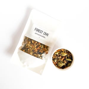 May include: A white pouch labelled FOREST CHAI containing a loose herbal tea blend with visible spices, alongside a small white bowl filled with the same mixture of dried ingredients including cardamom pods, peppercorns, and bark pieces.