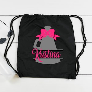 Puede incluir: Bolsa de cordón negra con un diseño de megáfono gris y un lazo rosa. El nombre "Kristina" está escrito en una pancarta blanca sobre el megáfono.