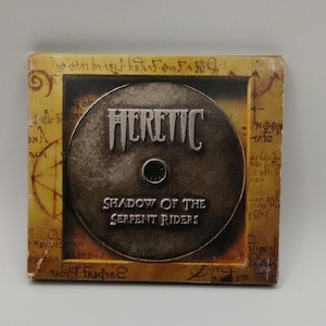 May include: A CD case featuring the game "Heretic: Shadow of the Serpent Riders." The disc is dark gray with the game title in silver. The case has a brown, aged appearance with script and designs.
