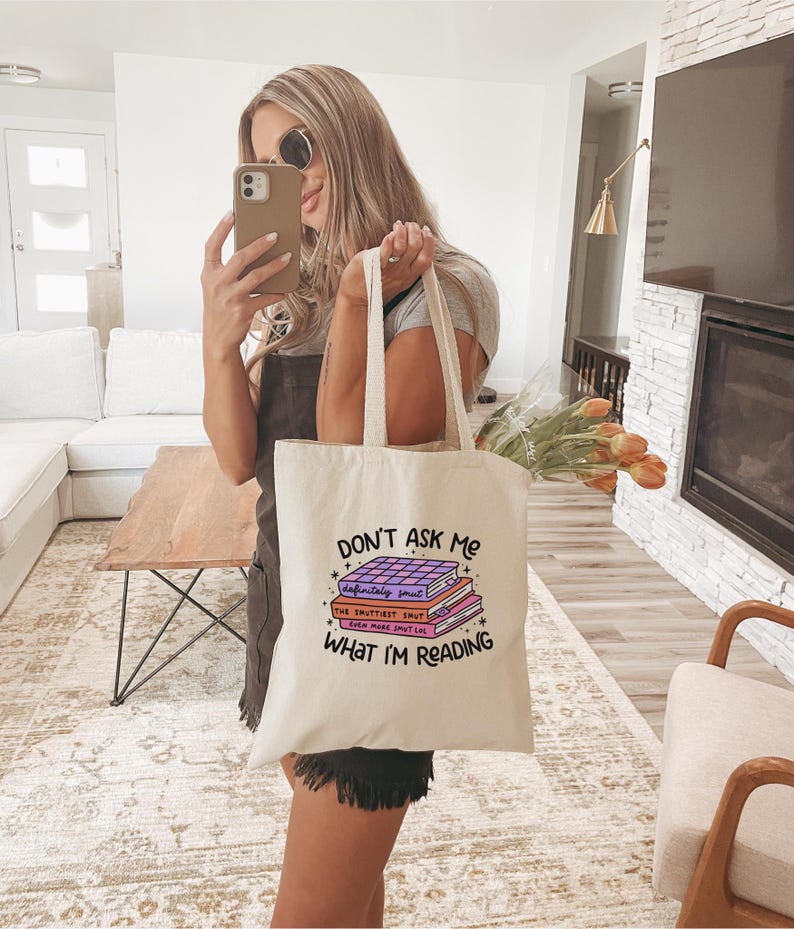May include: A white tote bag with the text "Don't Ask Me What I'm Reading" and a stack of books with the text "definitely smut", "The Sputtiest Smut", and "Even More Smut Lol" on the front.