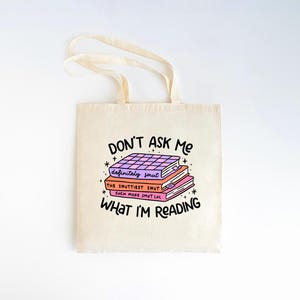 May include: A white canvas tote bag featuring a graphic of a stack of books with the text "DON'T ASK ME WHAT I'M READING." The books have titles that say "definitely smut", "THE SMUTTIEST SMUT", and "EVEN MORE SMUT LOL".