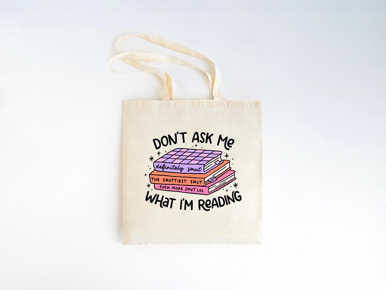May include: A white canvas tote bag featuring a graphic of a stack of books with the text "DON'T ASK ME WHAT I'M READING." The books have titles that say "definitely smut", "THE SMUTTIEST SMUT", and "EVEN MORE SMUT LOL".