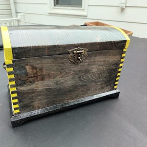 May include: A rectangular wooden chest with a dark stained finish and a curved lid. The edges feature bright yellow accents with visible finger joints. A decorative antique brass-colored metal latch is centered on the front of the chest.