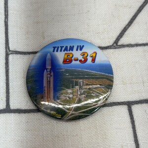 May include: A circular pinback button featuring a graphic of a Titan IV rocket on a launchpad. The text TITAN IV B-31 is printed in blue and orange lettering at the top. The background shows an aerial view of a coastal launch facility.