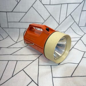 May include: A vintage orange plastic flashlight with a cream-colored bezel and a silver reflector. The handheld device features a top-mounted handle, a black power switch, and a metal loop for hanging, showcasing a classic utilitarian design.