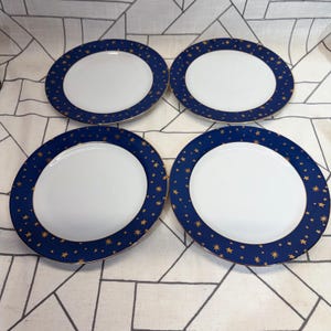 May include: A set of four round dinner plates, each featuring a solid white center and a wide, deep blue rim decorated with a pattern of small gold stars. The edges of the plates are finished with a thin gold trim, creating a consistent design.