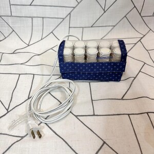 May include: A Remington electric hair setter featuring a rectangular blue fabric case with a white geometric pattern and a clear top. Inside are ten white cylindrical hair rollers arranged in two rows. A long white power cord with a plug is attached.
