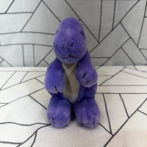 May include: A plush dinosaur toy featuring soft purple fur, a rounded head, and a light-colored cream belly panel. The stuffed animal is depicted in a seated position with short arms and legs, showcasing a simple, textured fabric design throughout.