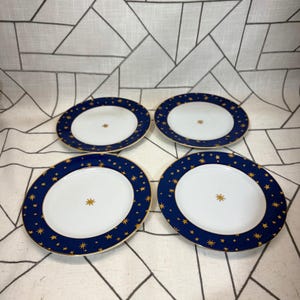 May include: A set of four round ceramic plates featuring a white center with a single gold star and a wide dark blue rim decorated with numerous gold stars of varying sizes. Each plate has a thin gold trim along the outer edge.