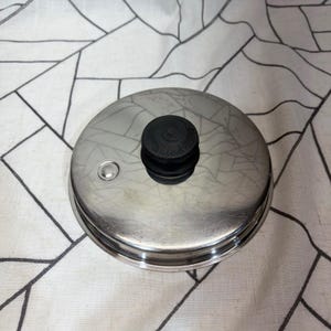 Könnte beinhalten: Ein runder Topfdeckel aus Edelstahl mit einer polierten, reflektierenden Oberfläche. Er verfügt über einen schwarzen Knopf in der Mitte mit dem eingeprägten Text Saladmaster und eine kleine runde Dampföffnung am Rand.