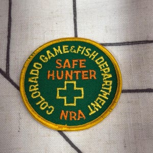 May include: A circular embroidered patch with a dark green background and a yellow border. The text reads COLORADO GAME & FISH DEPARTMENT, SAFE HUNTER, and NRA in yellow and orange. A yellow cross symbol is centered on the patch.