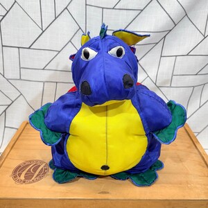 May include: A stuffed dragon toy with a blue head and body, yellow belly, and green accents on the wings and feet. The dragon has large white eyes, black nostrils, and a red patch on its back. The toy is sitting on a wooden surface.