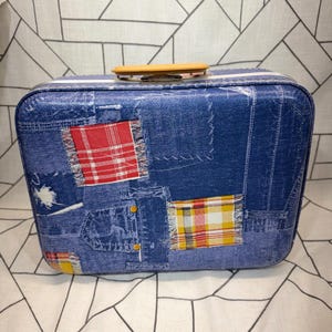 May include: A rectangular suitcase featuring a blue denim-look print with a patchwork design. The surface displays stitched fabric patterns, including red and yellow plaid squares, along with a tan plastic handle attached to the top edge.
