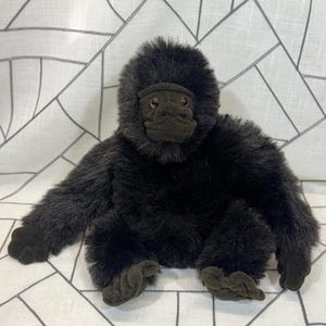 May include: A plush gorilla toy with dark brown fur and a detailed face. The stuffed animal has a seated pose with arms and legs extended. The gorilla's fur is soft and fluffy, with a realistic appearance.