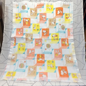 Puede incluir: Manta blanca de Snoopy con un patrón repetitivo de Snoopy en varias poses dentro de bloques geométricos coloridos. Los bloques son en tonos de naranja, amarillo y marrón, con acentos azules y blancos. La manta tiene un borde con volantes.