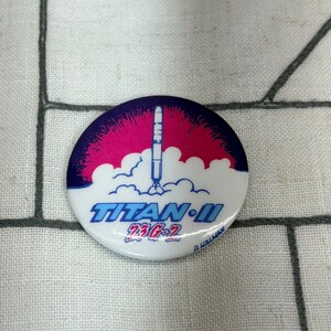 May include: A round pinback button featuring a graphic of a rocket launching into a dark purple and pink sky above white clouds. The text TITAN II and 23G-2 are printed in blue and red, with the name D. HEILMAN visible at the bottom edge.