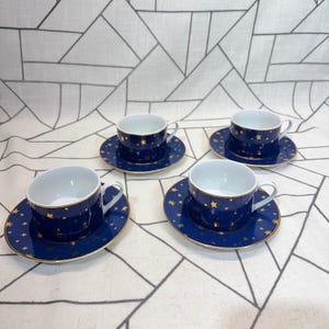 May include: A set of four dark blue ceramic teacups and matching saucers, each featuring a gold star pattern and a gold rim. The cups have a white interior and a curved handle, while the saucers are decorated with gold stars of varying sizes.