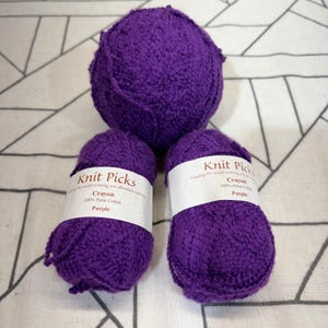 May include: Three balls of deep purple yarn made of 100% Pima cotton. Two balls feature white paper labels with the text Knit Picks, Crayon, 100% Pima Cotton, and Purple. The yarn has a textured, slightly nubby appearance throughout the surface.