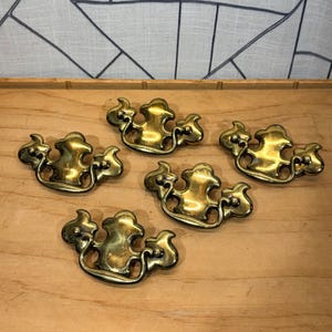 May include: Five antique brass drawer pulls with an ornate design. Each pull has a curved handle and stylized floral or cloud-like shapes. The pulls are arranged on a light brown wooden surface.