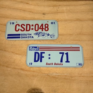 May include: Two vintage South Dakota license plates. One plate has "CSD*048" in red, and the other has "DF*71" in blue. Both plates feature the state name and a depiction of Mount Rushmore.