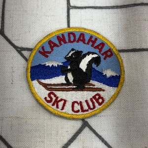 May include: A circular embroidered patch featuring a black and white skunk standing on brown skis against a light blue sky and white snowy mountain background. The text KANDAHAR SKI CLUB is stitched in red letters with a yellow border.