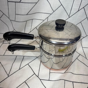 Könnte beinhalten: Doppelkochtopf aus Edelstahl mit Deckel und schwarzen Griffen. Der Topf hat einen kupferfarbenen Boden und einen schwarzen Knopf auf dem Deckel. Der Topf ist zum Dämpfen oder Schmelzen von Zutaten konzipiert.