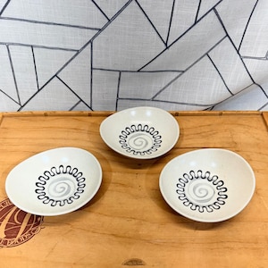 May include: Three off-white ceramic bowls with a black and gray spiral design in the center. The bowls are arranged on a wooden tray, with a patterned fabric in the background. The bowls have a unique, organic shape.