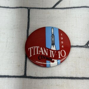 May include: A round pinback button featuring a red background with a vertical light blue stripe. A white illustration of a Titan IV rocket is centered on the stripe. The text reads TITAN IV 10 CENTAUR and the year 1994 in white and black.