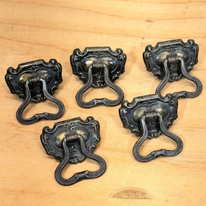 May include: Five antique bronze drawer pulls with ornate, oval backplates and curved handles. The hardware has a vintage aesthetic, with a dark, aged finish. The pulls are arranged on a light wood surface.