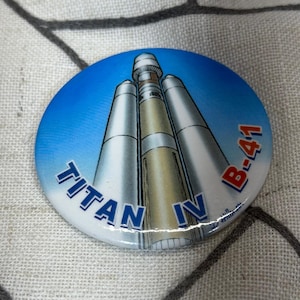 May include: A circular pinback button featuring a graphic illustration of a Titan IV rocket against a blue sky background. The text TITAN IV B-41 is printed in bold blue and red lettering across the lower portion of the button surface.