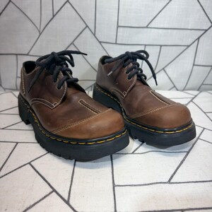 May include: A pair of brown leather oxford shoes featuring black laces, a thick black lug sole, and prominent yellow stitching along the welt. The shoes have a rounded toe box with a decorative seam detail across the top and a matte finish.