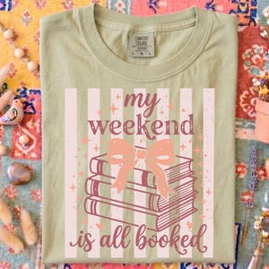 Op de afbeelding: Saliegroen t-shirt met de tekst "my weekend is all booked" in een speels lettertype. Het ontwerp toont een stapel boeken met een strik, tegen een gestreepte achtergrond. Het shirt heeft een ronde hals en een Comfort Colors label.