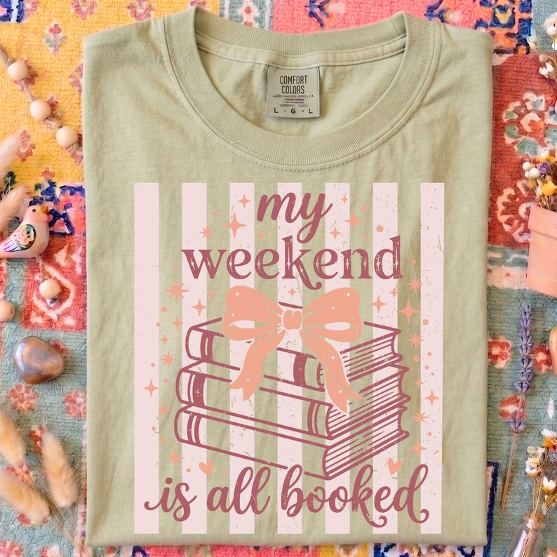 Op de afbeelding: Saliegroen t-shirt met de tekst "my weekend is all booked" in een speels lettertype. Het ontwerp toont een stapel boeken met een strik, tegen een gestreepte achtergrond. Het shirt heeft een ronde hals en een Comfort Colors label.