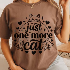 May include: A brown short-sleeved t-shirt featuring a black graphic print of three cats and the text "just one more cat" arranged in a circular design with small hearts and stars surrounding the lettering and the cat illustrations.