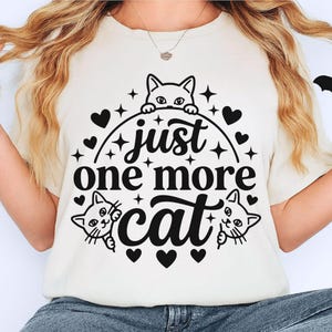 May include: A cream-coloured t-shirt featuring a black graphic print with the text "just one more cat" centred in the design. The illustration includes three cat faces peeking out from behind the lettering, surrounded by small hearts and stars.