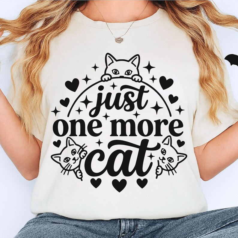 May include: A cream-coloured t-shirt featuring a black graphic print with the text "just one more cat" centred in the design. The illustration includes three cat faces peeking out from behind the lettering, surrounded by small hearts and stars.