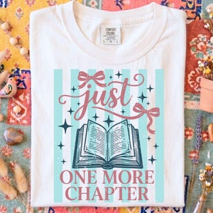 Op de afbeelding: Wit T-shirt met een grafisch ontwerp met de woorden "just" en "one more chapter" in roze en teal. Het ontwerp bevat een open boek, strikken en sterren. Het shirt heeft een Comfort Colors label.