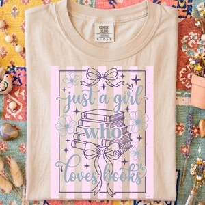 Op de afbeelding: Beige Comfort Colors T-shirt met de tekst "just a girl who loves books". Het ontwerp bevat een stapel boeken, strikken, bloemen en een gestreepte achtergrond in roze en paars.