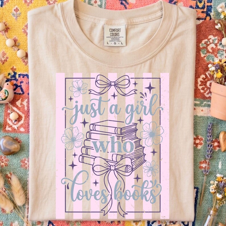 Op de afbeelding: Beige Comfort Colors T-shirt met de tekst "just a girl who loves books". Het ontwerp bevat een stapel boeken, strikken, bloemen en een gestreepte achtergrond in roze en paars.