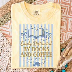 Op de afbeelding: Lichtgeel T-shirt met een blauw-wit gestreept ontwerp. Het shirt heeft een afbeelding van een open boek, strikken en een kopje koffie, met de tekst "Easily Distracted BY BOOKS AND COFFEE."