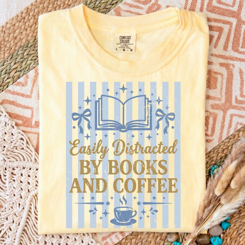 Op de afbeelding: Lichtgeel T-shirt met een blauw-wit gestreept ontwerp. Het shirt heeft een afbeelding van een open boek, strikken en een kopje koffie, met de tekst "Easily Distracted BY BOOKS AND COFFEE."